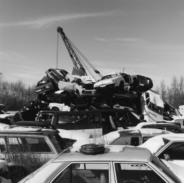Slightly elevated view of cars, vans, and trucks laying in a heap on top of each other, with a light cover of snow over them. A crane rises above the pile. 