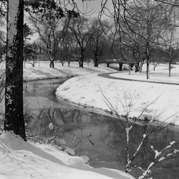 View from shoreline looking down at a light film of ice covering the surface of a river as it curves around a bend towards a bridge in the distance. A path and trees are winding along the bank on the opposite shoreline along the river. A house is in the far distance.