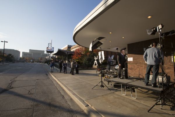 A camera operator is filming a reporter outside the Milwaukee Theater at the Republican presidential debate. Other television crews are setting up their equipment or are talking in small groups. A large line of people is standing under the awnings of the Milwaukee Theater waiting to be let inside. The Milwaukee County Courthouse is in the far distance at the end of the street.