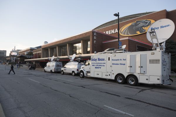 View down street towards three news vans sitting outside the UW-Milwaukee Panther Arena, a building adjacent to the Milwaukee Theater which is hosting the Republican presidential debate. A security guard is carrying a blockade across the street, and a police officer is standing next to a motorcycle. A line of men and women are waiting to be let inside the theater.