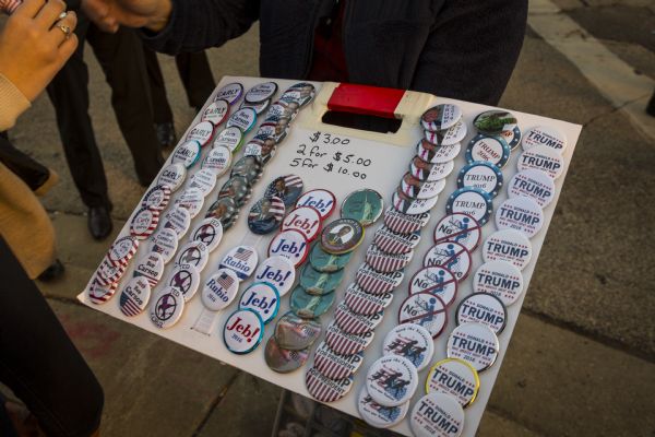 A woman is holding a board filled with campaign buttons for several Republicans running for President. Most of the buttons state the name of the candidate, but two sets display anti-immigration sentiments, particularly relating to the Mexican and American border. A small handwritten note on the board lists the price of the buttons as three dollars, two for five dollars, or five for ten dollars.