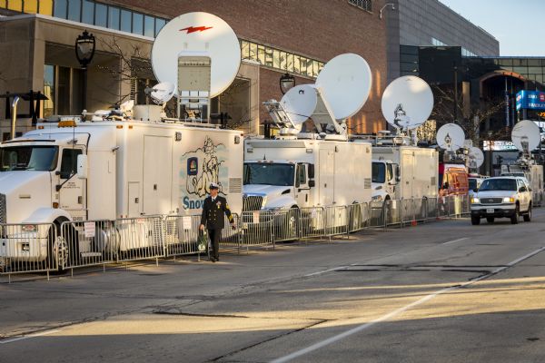 View across street towards a man in formal Navy dress walking down a line of metal fence barriers blocking off broadcasting vans from news stations parked near a building.