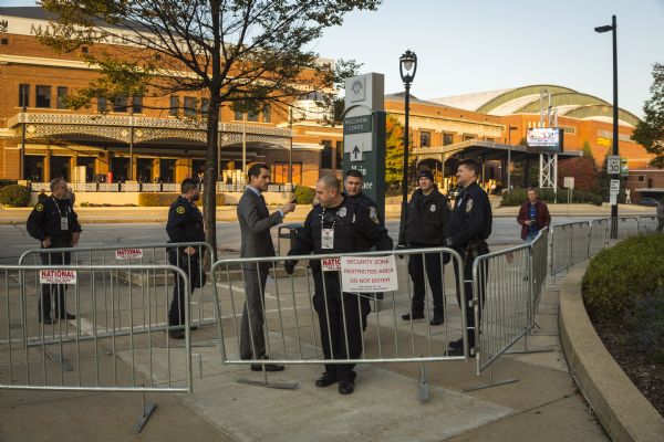 A police officer is setting up the metal fence barricade across the street from the Milwaukee Theater. Five other police officers are standing around him. A man in a suit is talking to the policeman setting up the barricade. Another man in a flannel jacket and shirt is approaching the group from behind and is carrying an SLR camera.
