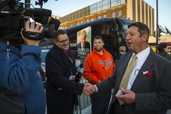 Michael Petyo is shaking hands with a reporter from 12 News, as a camera operator is filming them. Petyo is wearing a suit and a red flower pin. Another man in an Oklahoma State sweatshirt standing behind the two men and watching, is wearing the same pin as Petyo. A large black bus featuring an advertisement for Donald Trump's book "Crippled America" is in the background.