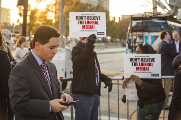 A man and a woman are facing each other and leaning against a metal barricade fence, each holding up signs from the Media Research Center that read: "Don't Believe the Liberal Media." A young man in a suit looking at his smart phone is in the foreground on the left. 