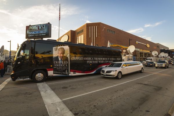 View across intersection towards a large black bus in front of the U.W. Milwaukee Panther Arena. On the bus is an advertisement for Donald Trump's book <i>Crippled America</i>. A white limousine is in the lane next to the bus. Media vans are in the background on the right.