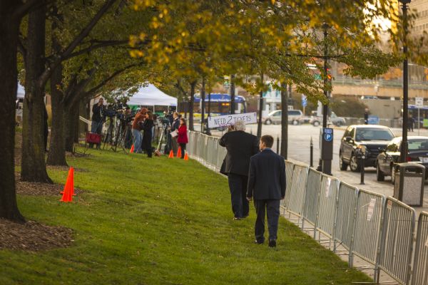 View down grassy lawn bordered by trees towards two men wearing suits  who are walking along a metal barricade blocking the sidewalk and street on the right. The older man is holding a handwritten sign reading: "Need 2 Tickets. Please Help." There is a group of reporters and camera operators near a tent in the background. 