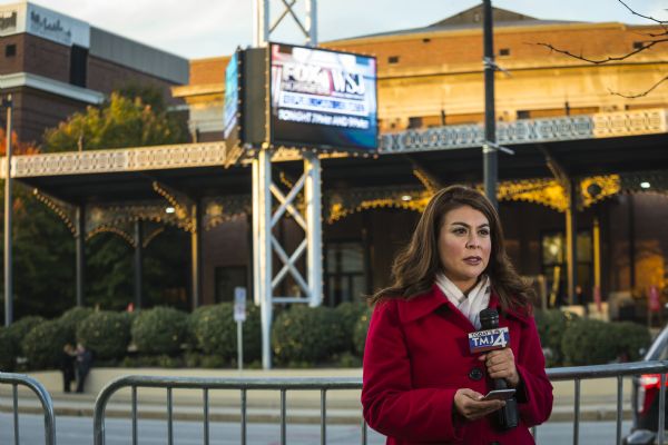 Veronica Macias, a reporter for TMJ4, standing across the street from the Milwaukee Theater. She is holding a microphone in one hand and a smart phone in the other. Behind her is a metal barricade.