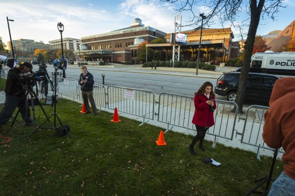 Slightly elevated view of two reporters, Veronica Macias from TMJ4, and a man from WFRV-TV Local 5, who are standing on grass behind metal fence barricades and looking at their smart phones as they are talking into microphones. Camera operators are filming and broadcasting their reports. Across the street is the Milwaukee Theater, which is hosting the Republican presidential debate.