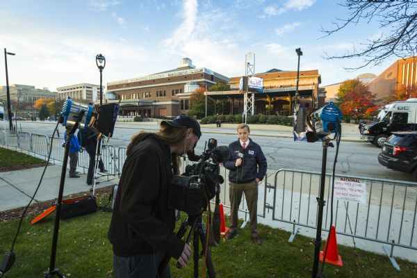 A young male reporter for WFRV-TV Local 5 is holding a microphone and talking to the camera, which is operated by a camera operator standing in the foreground. Across the street is the Milwaukee Theater, which is hosting the Republican presidential debate.