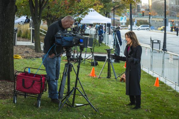 A female reporter and a male camera operator are standing on a grassy strip near metal fence barriers blocking off a street on the right. The reporter is gesturing with her hands while reporting on the Republican presidential debate. Other television news crews are setting up in the background.