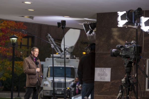 A reporter is standing outdoors under the awning of a building holding a microphone. A camera operator is aiming lights and a camera at him. A sign on the wall behind the camera operator reads: "FBN Outside Cam." In the background is a media van. 