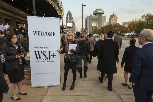 Two women are standing on either side of a large welcome sign from "The Wall Street Journal." The woman on the left is holding a phone in her hand and is wearing a staff badge for the Republican presidential debate. The woman on the right is holding a phone in one hand and a folder and pen in the other. A line of people are walking past them into the Milwaukee Theater which is hosting the debate.