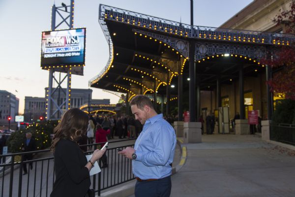 A man and a woman are standing and facing each other, but looking down at their smart phones. Behind them, lines of people are wait to enter the Milwaukee Theater for the Republican presidential debate.