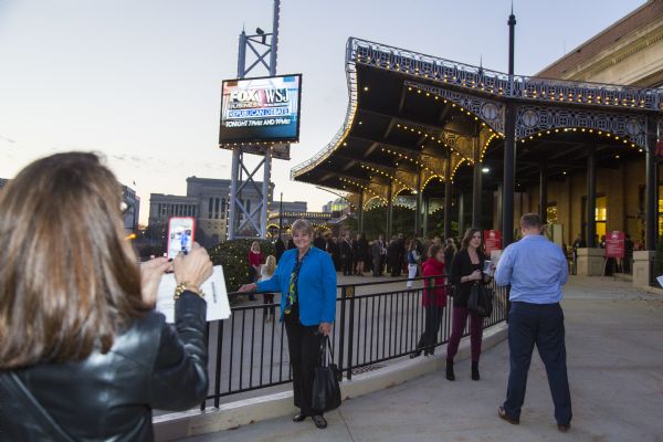 A woman in a black coat is taking a picture with her phone of another woman in a blue blazer, who is standing in front of a metal barrier fence. They are in front of the electronic sign announcing the Republican presidential debate, outside of the Milwaukee Theater.