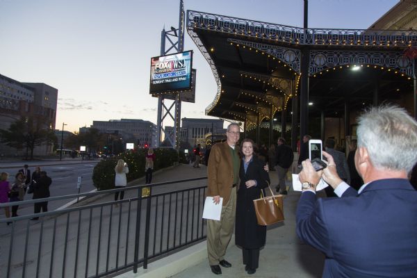 A couple poseing outside the Milwaukee Theater under the electronic sign announcing the Republican presidential debate. Another man, holding two phones, is taking their picture.