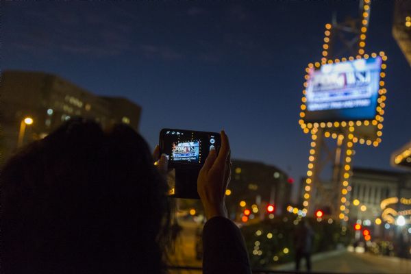 View from behind of a woman holding up her smart phone to take a photograph of the electronic sign outside of the Milwaukee Theater. The sign is advertising the Republican presidential debate, hosted by Fox Business News and "The Wall Street Journal." Lights illuminate the large screen against the darkening sky.