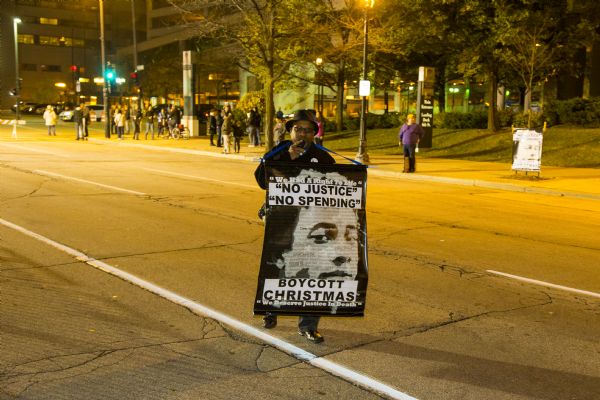 View towards a man walking alone down the middle of the closed street outside of the Republican presidential debate at the Milwaukee Theater. The man is holding a microphone or voice amplifier up to his mouth and is carrying a large sign hanging from his neck. The sign bears a distorted black and white image of a man covered with the names of African-American people who have been shot by cops or vigilantes, such as Corey Stingley and Trayvon Martin. The sign reads: "We had a right to life," "No Justice, No Spending," "Boycott Christmas," and "We Deserve Justice in Death." There is a small crowed of people on the opposite side of the street.