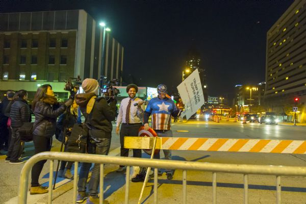 Two women are operating cameras recording a reporter who is holding a microphone up to another man, Todd Eiler, dressed as Captain America. Todd holds a sign reading: "Trump is Our Super Hero. Mr. Trump Please Sign My Shield." They are standing outside of the blocked off street leading to the Republican presidential debate at the Milwaukee Theater.