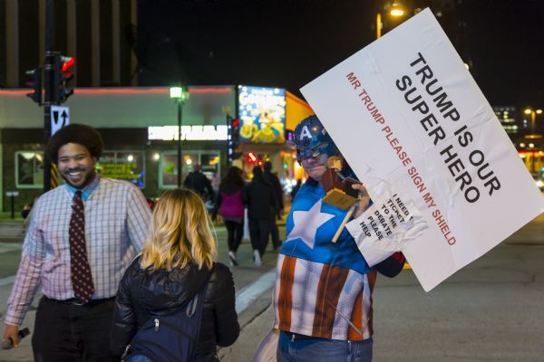 Todd Eiler, dressed as Captain America, holds up a sign reading: "Trump is Our Superhero. Mr. Trump Please Sign My Shield." Attached to that sign is a smaller one reading: "I need 1 ticket to the debate, please help." He is holding a small figurine of Donald Trump sitting at a desk. Next to him is comedian Tim Barnes holding a microphone. In the foreground is a young woman walking in the opposite direction.