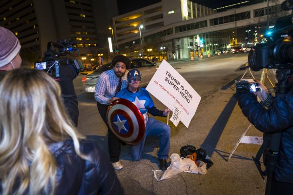 Todd Eiler, dressed as Captain America, kneels and poses on one knee on the ground, next to a small pile of plastic bags. He is holding a sign reading: "Trump is Our Superhero. Mr. Trump Please Sign My Shield." Another man, comedian Tim Barnes, is posing behind the costumed man, holding onto his back and looking at the cameras. Men and women are filming the scene with professional cameras and a smartphone.