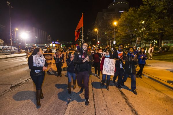 View of the front of a line of protesters marching down the street near to the Republican presidential debate at the Milwaukee Theater. The group is clapping and shouting as they march, and are carrying flags, signs, and banners. One of the women in front is holding a sign that reads: "Black Lives Matter."