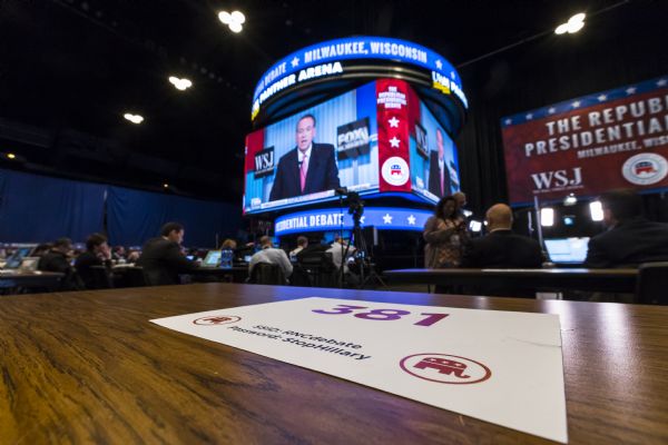 View across table with a sign indicating a seat number. In the background, reporters and journalists are sitting at tables with their equipment inside the Panther arena as they watch the Republican presidential debate on a large three-sided screen. Mike Huckabee is on the screen in the background. The sign on the table has the Republican elephant, and reads: "381. SSID: RNCdebate. Password: StopHillary."