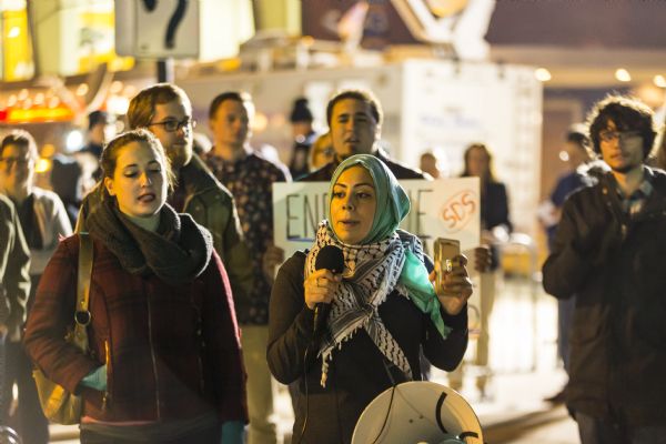 A young woman wearing a hijab is talking into a microphone while talking to a crowd of young men and women. A megaphone is strapped to her waist. 