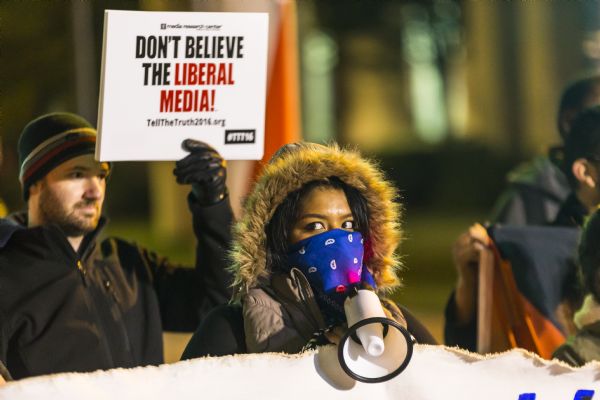 A young woman is holding up a sign for the Student Union Movement, a social justice group based out of Milwaukee Area Technical College, and a megaphone. She is wearing a hood and blue bandana to cover her face. Behind the woman is a young man who is holding up a sign reading: "Don't Believe the Liberal Media," a slogan for the Media Research Center.