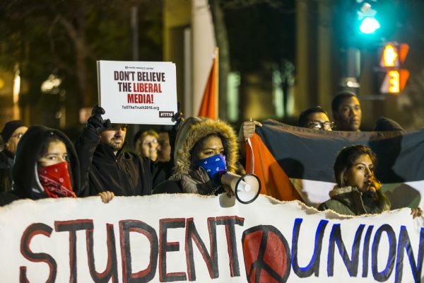 Three women are holding up a banner for the Student Union Movement, a social justice group based out of Milwaukee Area Technical College. Two of the women are wearing hoods and bandanas to cover their faces, and the woman in the middle is speaking through a megaphone. A man directly behind them is holding up a counter protest sign reading: "Don't Believe the Liberal Media," a slogan for the Media Research Center. Another young man on the right is holding a Palestinian flag.