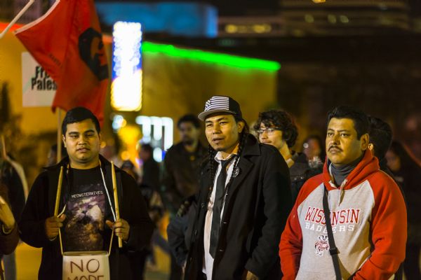 Three men are standing amongst the protesters outside the Republican presidential  debate. The man on the right is wearing a Wisconsin Badgers sweatshirt. The man in the middle is wearing a coat and tie, along with a baseball cap with a black and white image of the American flag. The third man on the right is wearing a small drum hanging on a strap around his neck, and and is holding up a drumstick. A red flag with the image of Che Guevara is in the background.