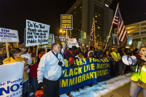 A young woman on the right is walking in front of a large crowd of protesters. She is wearing a reflective vest and is speaking through a megaphone. The crowd is holding up flags, banners, and posters. The signs read: "Black Lives Matter, Immigrant Justice, Fight for $15." One poster bears the image of Our Lady of Guadalupe. Along the far left an African American woman is holding a handwritten sign that reads: "Do you think people who work full time should have to rely on public assistance??" 