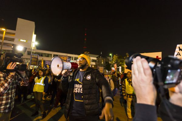 A young African American man is standing in the middle of a crowd speaking through a megaphone. Two camera operators are filming him as he is speaking. He is wearing a "Fight for $15 Wisconsin" shirt under a coat, and a packers knit hat. The crowd around him is holding up signs reading: "Black Lives Matter, Immigrant Justice, and Fight for $15." 