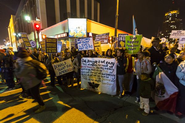 A large crowd of protesters standing on the street outside the Republican presidential debate. The group are mostly college age, although a young boy is standing in the front of the crowd. The protesters are holding signs, most reading: "Black Lives Matter, Immigrant Justice, and Fight for $15," but a few people are holding handmade signs such as: "On Land of Native Americans All Are Immigrants" and "Donald Trump for Amerikkka." A student group, MEChA de UW-Madison, is holding up a large banner which reads: "Our Struggle Is Not Easy. Those who oppose our cause are rich & powerful and they have many allies in high places. We are poor, our allies are few but we have something the rich do not own. We have our bodies & spirits & the justice our cause as our weapon — Ceasar Chavez." 
