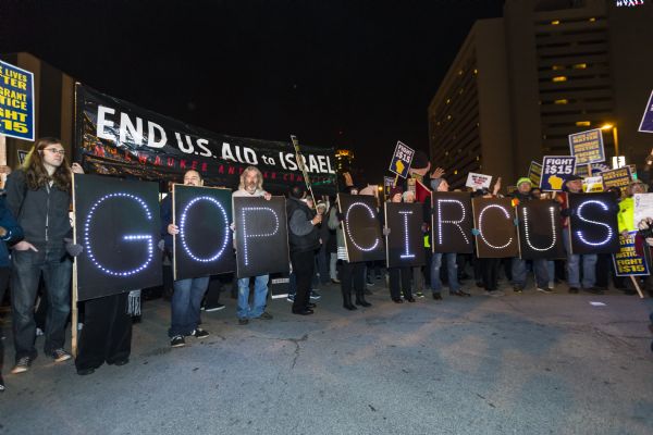 Members of the Overpass Light Brigade, a Wisconsin activist group, are standing in a semicircle holding up boards painted black with letters formed by lights. Together the letters spell the words: "GOP Circus." Other protesters are standing around and behind the group holding their own signs calling for a higher minimum wage, immigrant rights, and racial equality. Two people behind the Overpass Light Brigade are holding up a banner reading: "End U.S. Aid to Israel. Milwaukee Anti-War Committee."