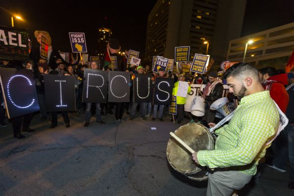 A man is standing in the foreground on the right, near a group of protesters seen in the background. The man is holding a drum slung over his right shoulder, a drum stick in his left hand, and a smart phone in his right. Behind him another man is beating on a plastic tub with two drumsticks. In the background the Overpass Light Brigade is forming a semicircle with their signs, reading: "GOP Circus." Other people in the crowd are holding up signs reading: "Black Lives Matter, Immigrant Justice, and Fight for $15." Two large puppets of Donald Trump and Scott Walker are in the crowd.