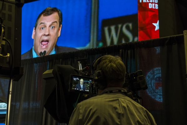 View from behind of a camera operator filming a reporter, Ali Velshi, in the press staging area in the Panther Arena during the Republican presidential debates. Velshi is on the small screen of the camera. Governor Chris Christie is on the large screen in the background just above a curtain.