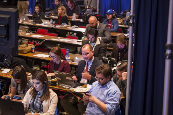 View looking down at reporters and journalists sitting at long tables, typing on their computers or watching the Republican presidential debate. One of the journalists, Scott Bauer, a correspondent with "The Associated Press," is looking up and giving a "thumbs up."