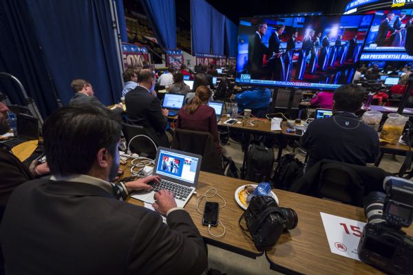 View over man's shoulder as he is editing and recording footage of the Republican presidential debates on his computer, with his smart phone and two large cameras sitting on the table next to him. Other journalists around him are working on their computers or writing in notebooks while watching the large screens televising the debate. Some of the presidential candidates are on the screen standing at their podiums.