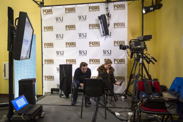 Two reporters are sitting and waiting on a small stage in the spin room for the nominees to emerge from the Republican presidential debate. They are looking at their phones and a screen set up on a chair. Video cameras, screens, computers and sound equipment are around the stage. A backdrop screen has the logos of Fox Business News and The Wall Street Journal, the sponsors of the debates.