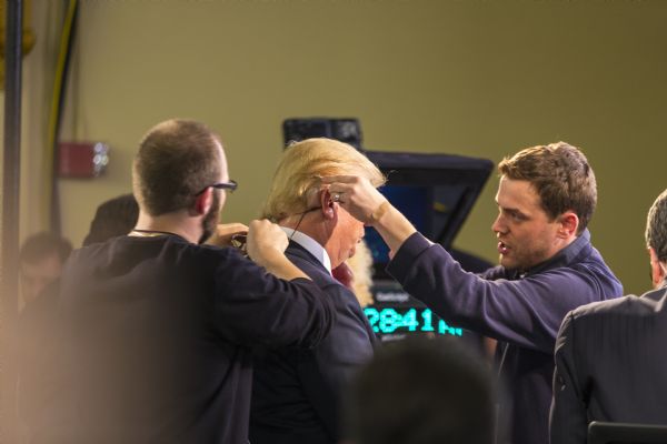 Two men are attaching a small microphone to Donald Trumps ear and fix ing the wire to his back. Trump is sitting on a stage next to a reporter in the spin room, preparing for his interview.
