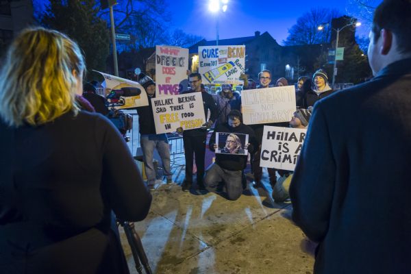 A group of anti-Hillary protesters posing outdoors for journalists and photographers with their handmade signs. In the foreground a woman stands behind a camera on a tripod on the left, and a man stands along the right.
