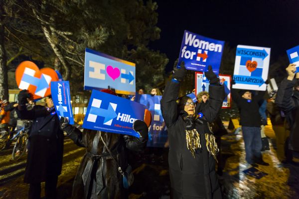 Two middle-aged women are holding up signs in support of Hillary Clinton. They are standing in front of a crowd of men and young women.