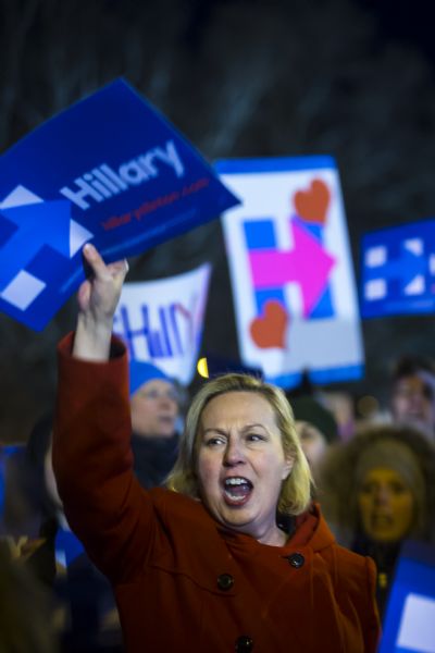 A woman in a red coat is holding up a sign in support of Hillary Clinton. Behind her is a crowd of Hillary supporters.