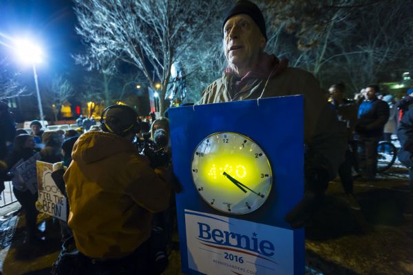 An older man is standing outdoors holding up a blue painted sign strapped around his neck. Attached to the sign is an analog clock, and a poster in support of Bernie Sanders. Behind him a photographer or camera operator is filming a group of young men and women holding their Bernie Sanders signs.
