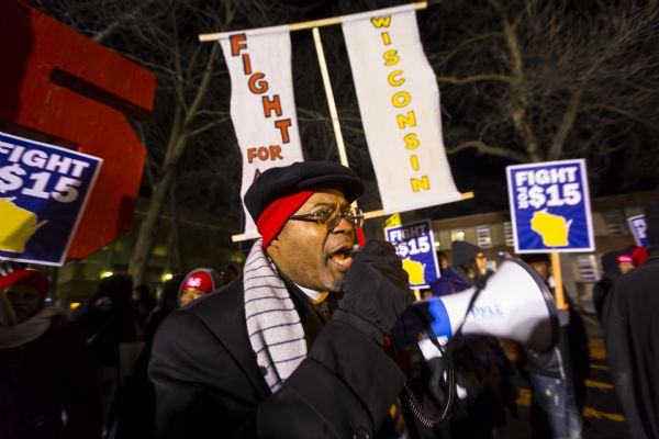 An African American man is standing outdoors speaking through a megaphone in front of a crowd in support of the fifteen dollar minimum wage.