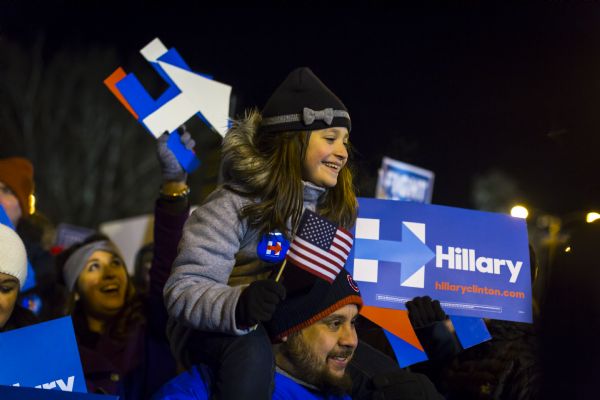 A father is carrying his young daughter on his shoulders. She is wearing a Hillary sticker on her coat and is waving a small American flag. A crowd of Hillary supporters is standing behind them holding up signs.