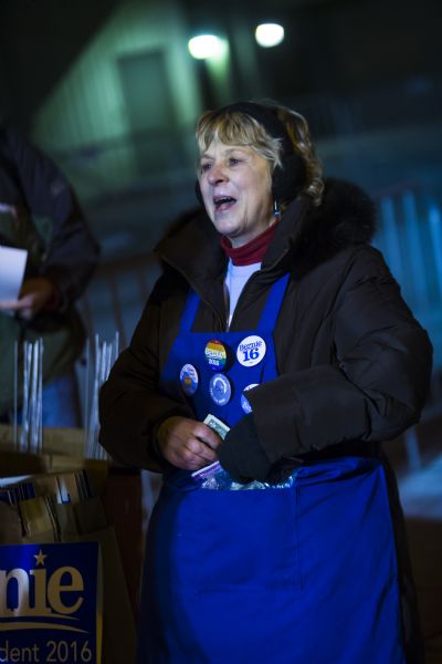A woman is standing and wearing an apron displaying political buttons in support of Bernie Sanders. The buttons are for sale, and she has money in the pocket of the apron. Next to her is a pile of Bernie Sanders lawn signs which are also for sale.
