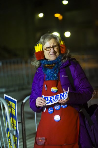 A woman is standing and holding Bernie Sanders bumper stickers while wearing Bernie political buttons on her apron that are for sale. She is wearing earmuffs in the shape of red containers holding french fries. The sign next to her reads: "Black Lives Matter, Immigrant Justice, Fight For $15."