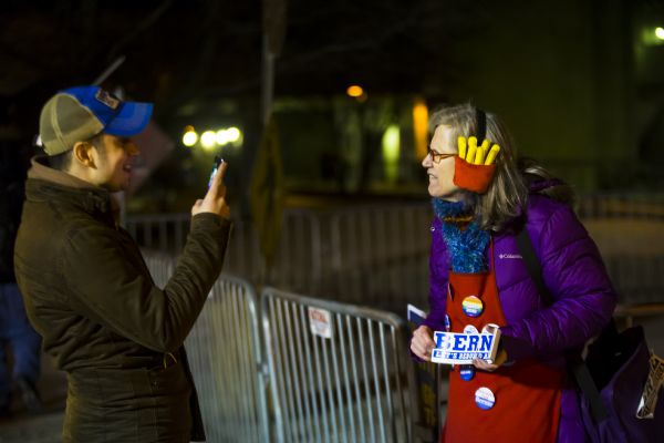 A woman selling Bernie Sanders political buttons and bumper stickers is posing for a young man who is taking a picture of her on his smart phone. She is wearing a red apron displaying the buttons, and earmuffs in the shape of french fries in red containers. 